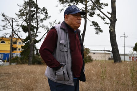 Man wearing collared long-sleeved shirt, sleeveless vest and baseball cap walks outside with hands in pockets.