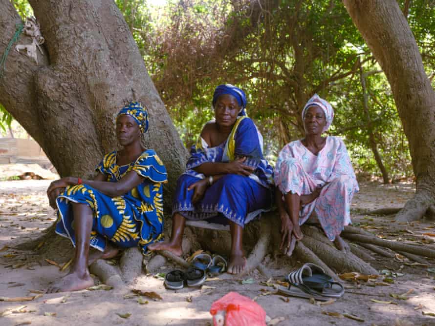 Three women sit at the foot of a tree, at the entrance to a sacred forest in Casamance, where residents come for medical treatments.