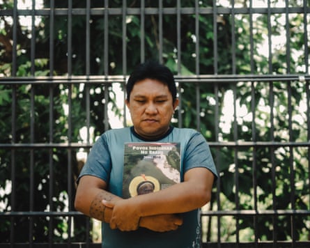 Dário Kopenawa crosses his arms holding a book Indigenous Peoples of Brazil, standing in front of a metal gate