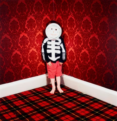 A child in red shorts, and wearing a black top with white lines to make it look like a skeleton, has in front of its face a circle of paper with a child’s face drawn on it. The child is standing on a red tartan-patterned carpet and the walls behind are papered in red flock wallpaper