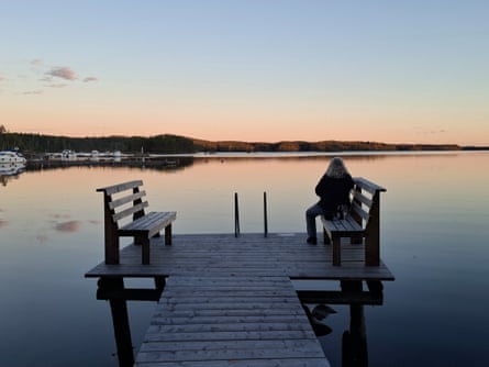 A blond woman sitting at the end of a wooden pier looking on to calm waters at sunset