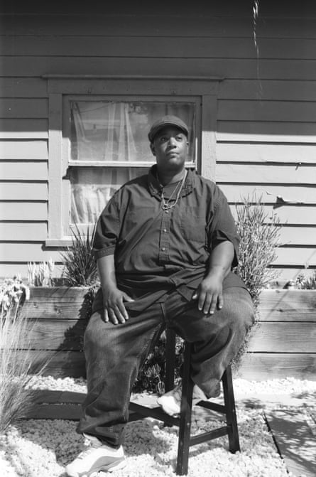 Malkia Cyril sits on a wooden chair outside her home, which has slatted wooden pannelling, in the sunshine.