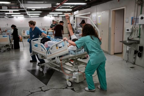 A equipe médica vestida de uniforme fica de costas para a câmera e empurra uma cama de hospital.