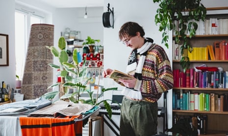 Wearing a jumper and scarf, Schranz stands reading a book in his light and airy apartment, with clutter and a colourful book shelf in the background.