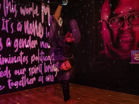 A young African-Caribbean man doing an acrobatic dance move in a dark room with painted words on one wall and a portrait of a young African-Caribbean man on the other.