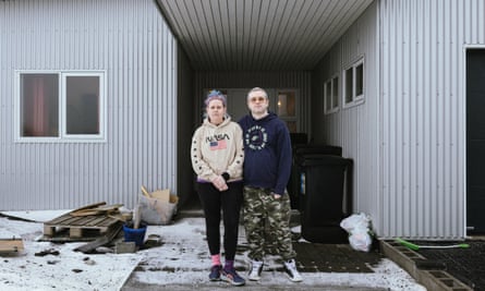 A woman with blue and purple hair tied up, a Nasa hoodie, leggings and trainers, and a man in a hoodie and combat trousers, stand by dustbins on a snowy path outside a house made of corrugated metal