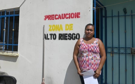 A woman stands in front of a building stamped with the words ‘Precaucion! Zona de Alto Riesgo’