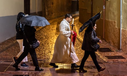 A man in a white robe walks along a wet street alongside women holding umbrellas