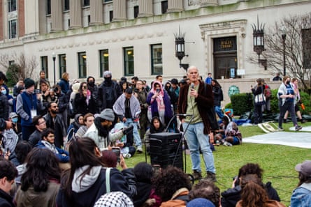 a man in jeans holds a microphone as people sit or stand nearby and look at him