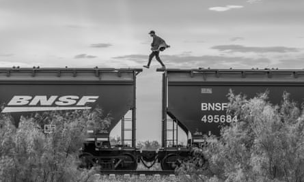 Black and white photo of a man attempting to step across the gap between two cargo wagons of a freight train in an empty landscape