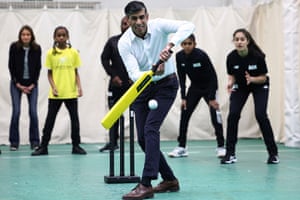 Rishi Sunak strikes a cricket ball with a bat in a practice net as young people stand behind in the slips