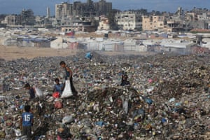 Children holding bags move over a huge mound of rubbish with a camp in the distance
