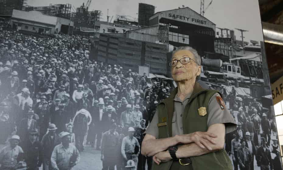 Betty Reid Soskin, a 94-year-old Black woman wearing a ranger vest and shirt stands for a portrait against an enlarged black-and-white second world war photo.