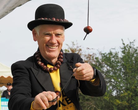 Man in a bowler hat with a ring of conkers on the brim, wearing a necklace made of conkers, aims a conker at another conker