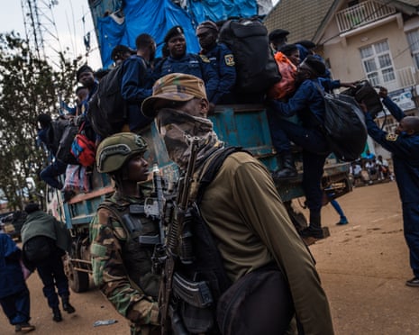 Two M23 rebels with rifles stand in front of a open-backed truck in which the police officers are sat