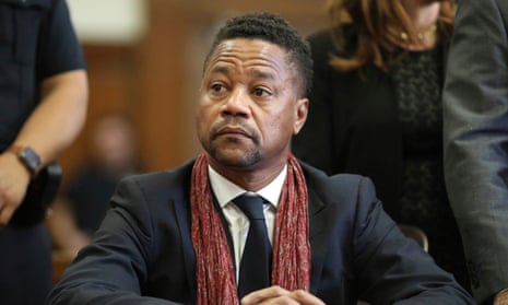 A Black man with trim hair a bit long on top, in black suit with red scarf, sits with hands crossed on table in front of him, looking serious and/or anxious toward someone on his right.