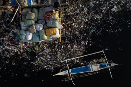 A boy sleeps on sacks of rubbish on a littered bank of the river beside a small makeshift canoe.