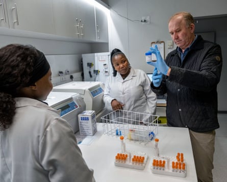 A doctor holds a sample pot over a table of vials in a laboratory, while two researchers in white lab coats look on