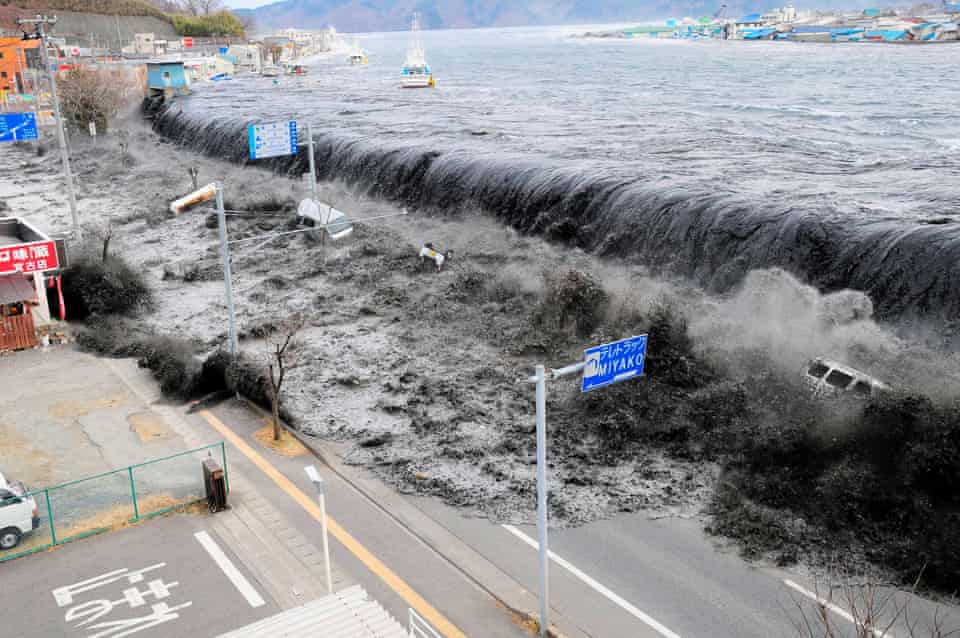 A huge wave looms over the oceanfront highway, sweeping cars along