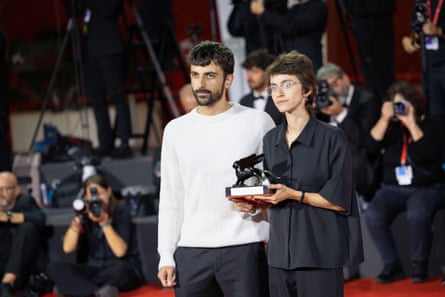 Yuval Abraham and Rachel Szor stand with their trophy in front of a bank of photographers. Abraham wears a white jumper and Szor a loose black shirt; they both look sombre. They both have short, dark hair and are in their early 30s; Abraham has a beard and Szor wears glasses.
