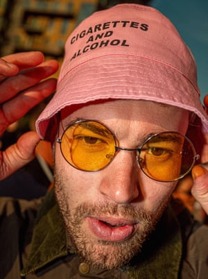 A young man wearing round tinted sunglasses and a pink bucket hat with ‘Cigarettes and Alcohol’ written on it.