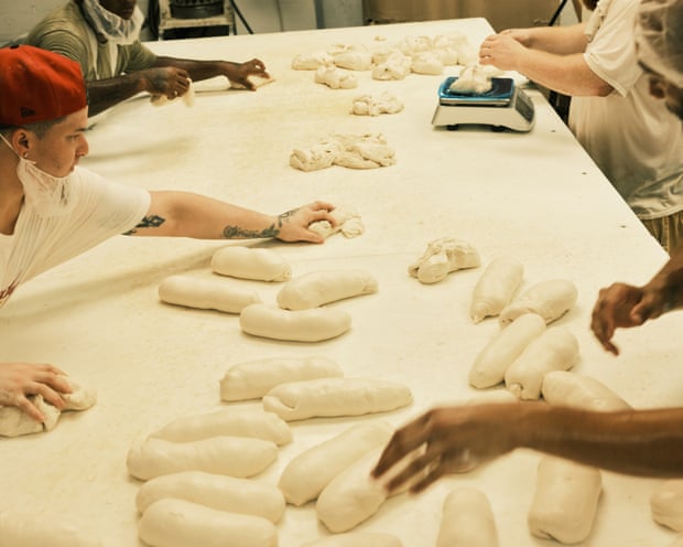 Four people work with dough at a long table.  Above right, one person measures lumps of dough on a scale, the person above left moves the lumps across the table, and two people below grab the lumps and shape them into loaves.