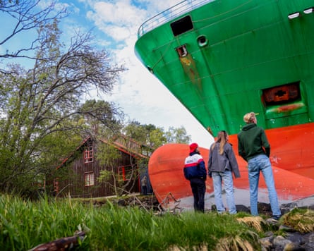 A closer view of the container ship with a house in the background.