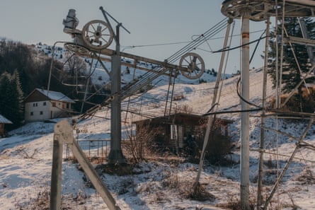 Old pylons and cables in front of a wooden cabin on a hillside with a dusting of snow over tussocks of grass and an alpine chalet in the background