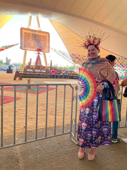 Latino man smiling wearing lipstick, wearing dress with dia de los muertos skulls, pink heels, scarf, flowered headdress.