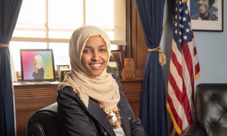 Ilhan Omar sitting at her desk in her office in Longworth House, Washington DC, a large window with curtain ties and a US flag behind her