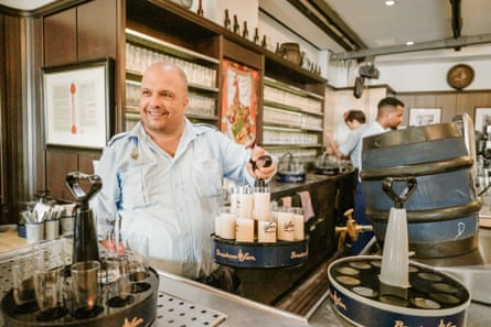 A barman standing behind a bar with barrels and glasses
