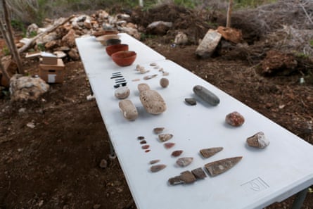 Tools, vases and pots are laid out on a white table in an outdoor area filled with dirt and dead vegetation.