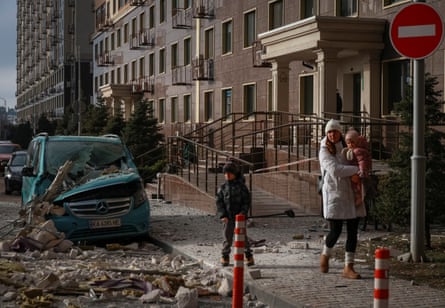 A woman and two children walk past a car crushed by falling masonry