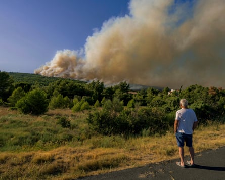 A man in shorts watches smoke from a fire filling the skyline over woodlands