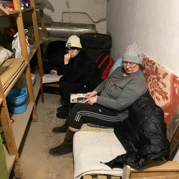 Veronika and her grandmother sit on a sofa in the basement of her grandparents’ home in Mariupol, both wearing woolly hats.