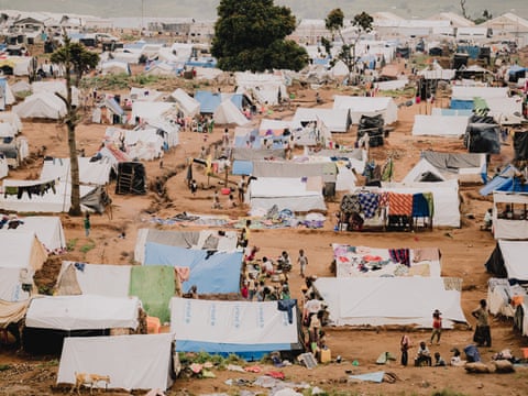 A view of tents pitched on muddy soil, trees and people walking around