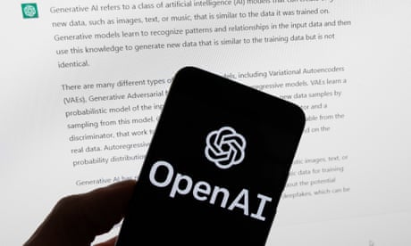 The OpenAI logo is seen on a mobile phone in front of a computer screen displaying output from ChatGPT