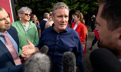 Keir Starmer in a blue shirt speaking to journalists holding microphones