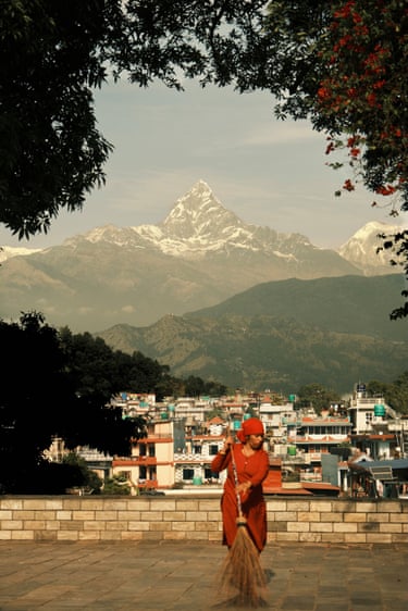 A woman in red sweeping a street in front of a city and mountain range
