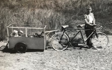 A black and white photo shows a woman with her hands on the handlebars of a tandem while two children sit in a trailer on the back
