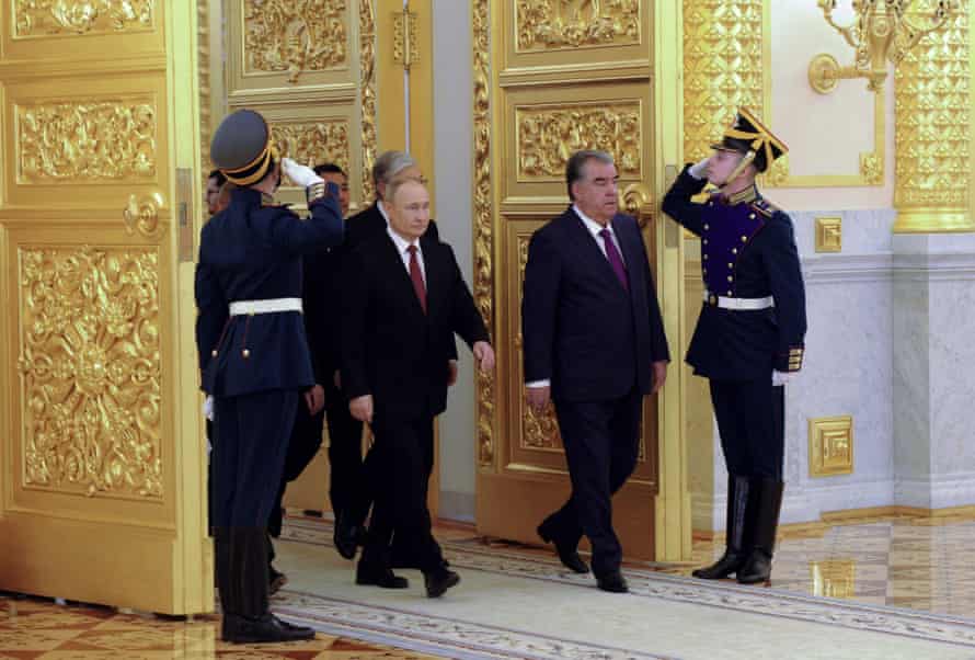 Guards in old-fashioned shako hats salute two men as they walk through huge golden doors