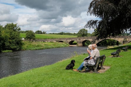 An older couple sitting on a bench eating ice-creams with a black dog sitting in front of them on the grassy banks of a river with an old stone bridge in the background