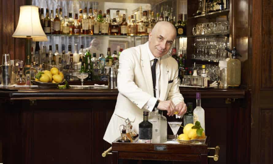 Besišypsantis vyras baltu švarku ir juodu kaklaraiščiu ruošia martinį vežimėlyje priešais barą