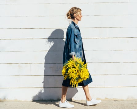 A person with blond hair, wearing an oversize demin jacket and white trainers walks in front of a white brick wall carrying a large bunch of yellow flowers by her side.