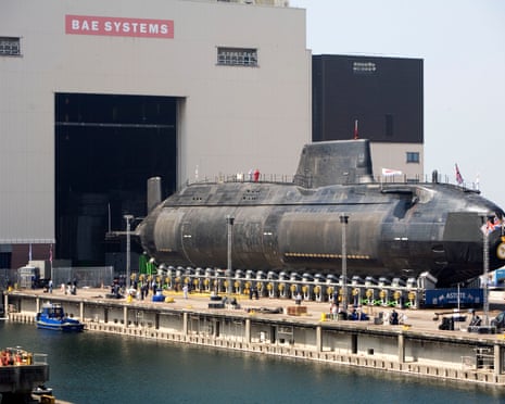 HMS Astute submarine sits on supports at a waterfront dock outside a BAE Systems building