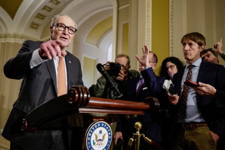 Chuck Schumer points while standing at a lectern as a crowd of journalists stands nearby recording him with their phones