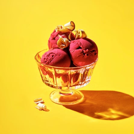 Three scoops of raspberry ice-cream in a stemmed dessert dish with shavings of wood sprinkled on top and a couple next to the base of the dish, against a bright-yellow background