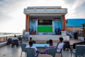 People sitting on chairs watch a televised football match on a screen on a terrace overlooking a beach