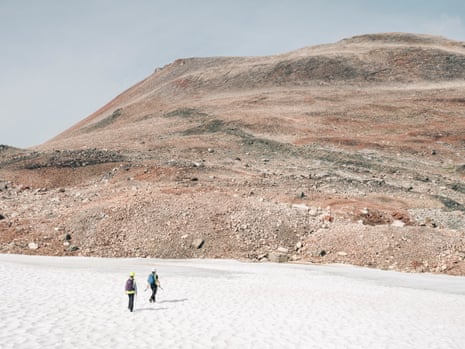 two people walk across snow towards brownish red terrain