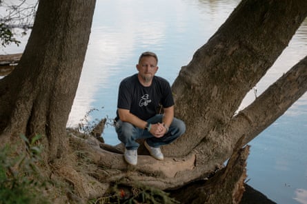 A man in a black T-shirt and jeans, sitting on a tree trunk with a lake behind him